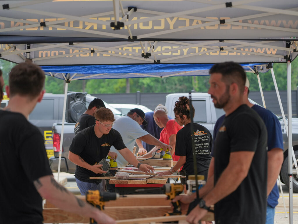 Impact Day 2026: Building More Than Beds 3 Legacy Construction crew members beginning the bed build at Legacy Day 2026 with lumber and power tools under a branded canopy tent