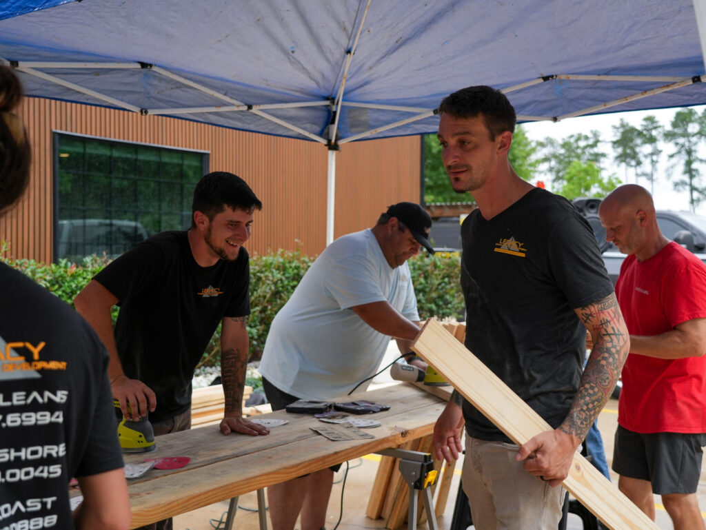 Impact Day 2026: Building More Than Beds 11 Volunteers sanding lumber boards under a canopy tent at Legacy Day 2026, a community bed build event supported by Legacy Construction and Development