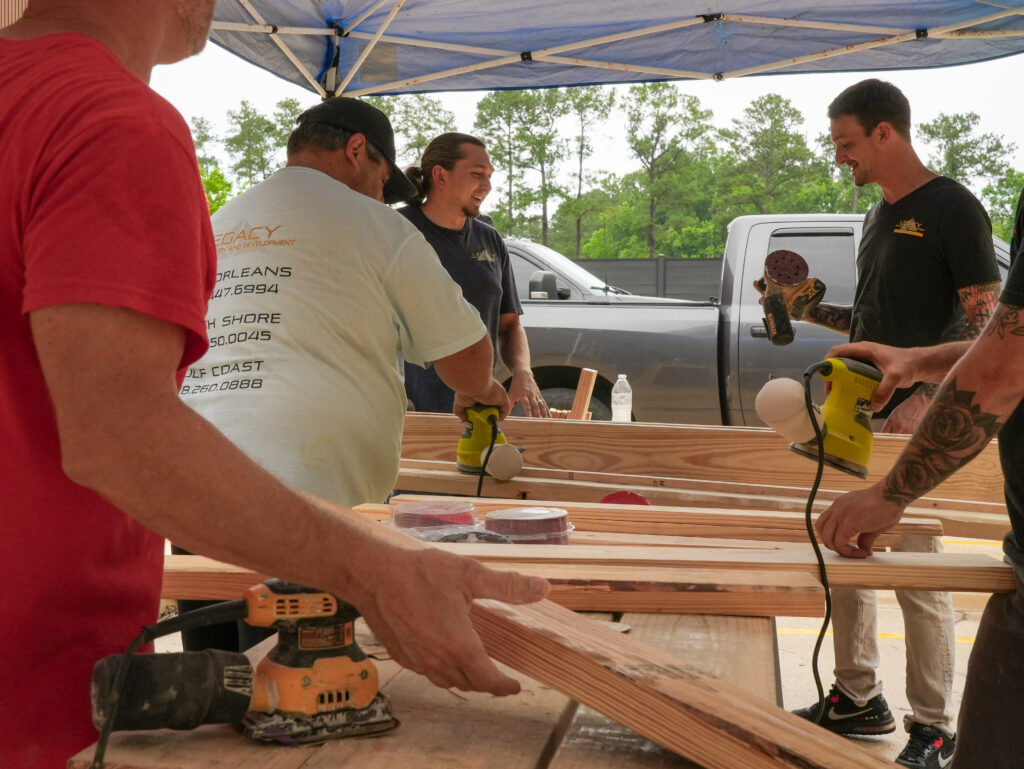 Impact Day 2026: Building More Than Beds 5 Volunteers and Legacy Construction employees assembling lumber under a canopy tent at the 2026 Legacy Day bed build event