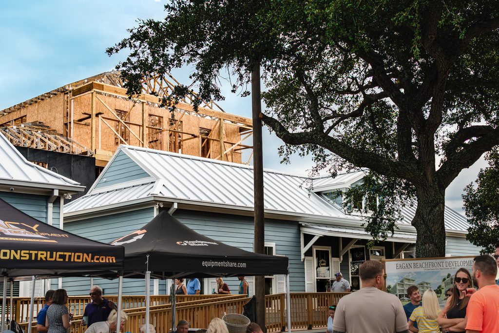 Guests mingle beneath an oak tree beside Eagan’s storefronts, with wood framing rising above the metal roofs.
